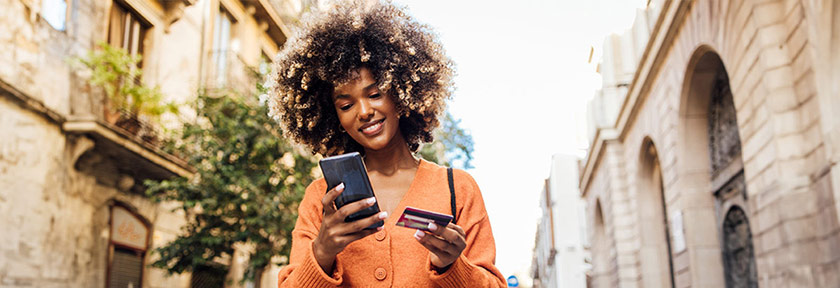 Woman outdoors holding a credit card in hand looking at her smartphone, mobile view, Nov 2023 blog, Travis CU,