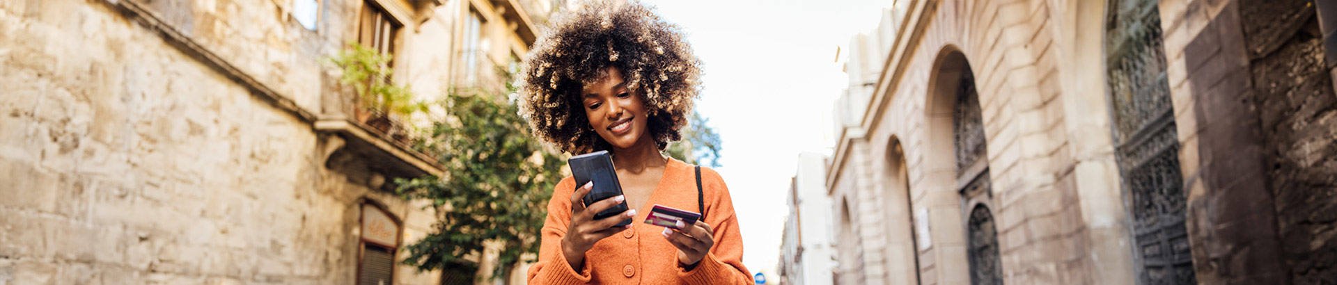 Woman outdoors holding a credit card in hand looking at her smartphone, Nov 2023 blog, Travis CU,