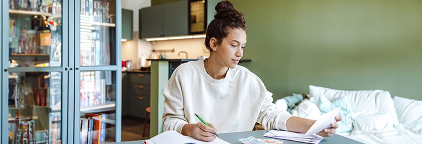 mobile photo of a woman sitting at a table in living room of a home paying bills, Aug 2023 blog, Travis CU,